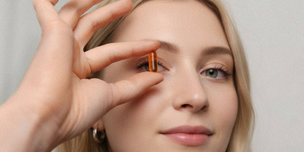 Nahaufnahme einer jungen blonden Frau, die eine rote Kapsel nu3 Curcuma Forte zwischen Daumen und Zeigefinger vor ihr Auge hält